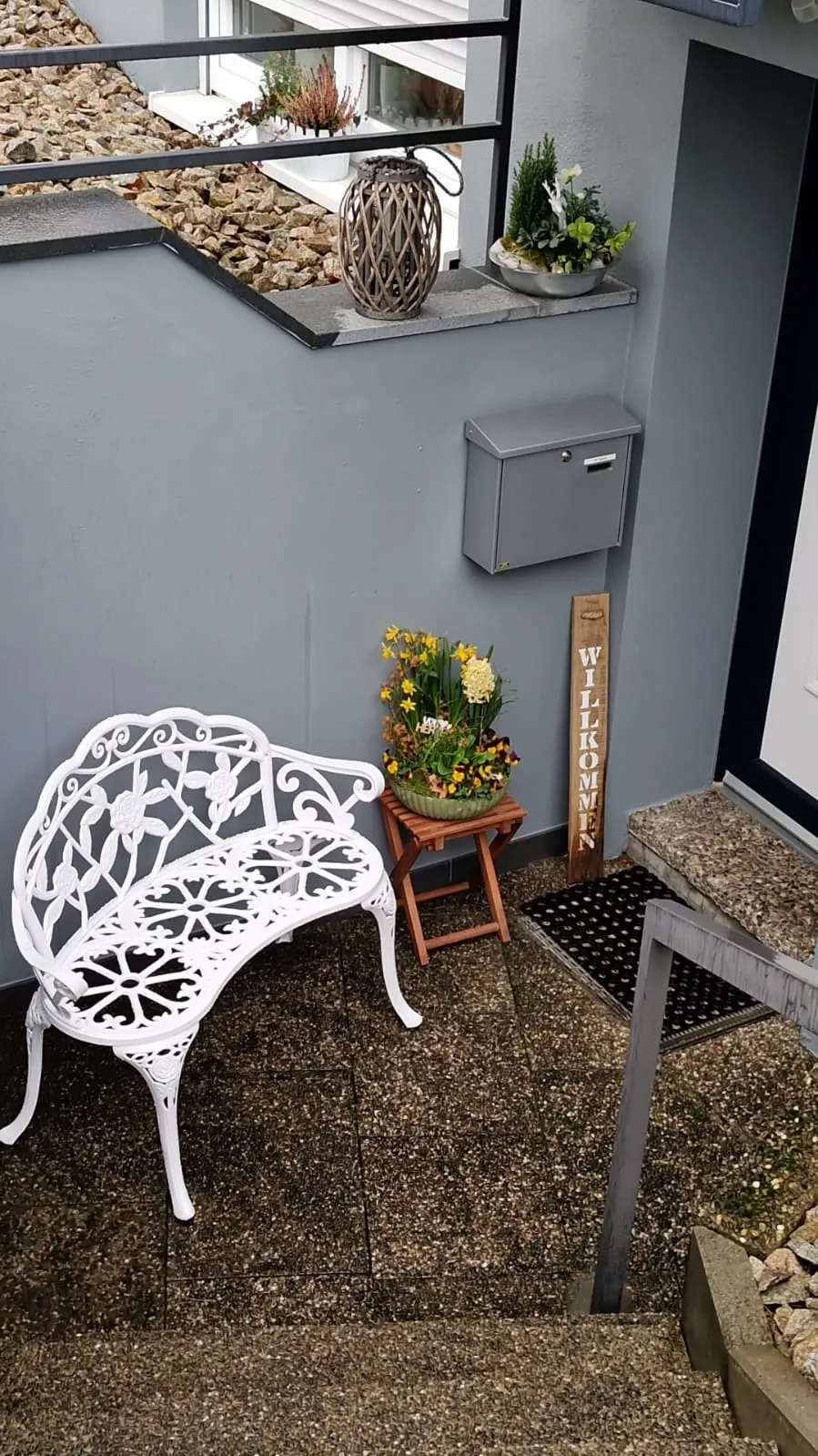 Eingangsbereich mit Willkommen-Schild und weißer Gartenbank vor dem Studio